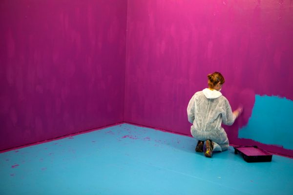 A young woman in protective overalls, wearing a safety mask, crouches in a bare blue room and paints the wall with bright pink paint.