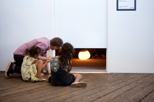 A man and two children crouch down to look through a low gap underneath a bare white door. Behind the gap is a young woman lying, cramped, next to a round glowing lamp.