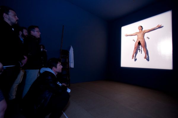 A naked woman sits, balancing, arms outstretched, on a bicycle seat, mounted high on the wall, in a glowing square of white light. Around 6 people watch the performer in the bare, darkened room.
