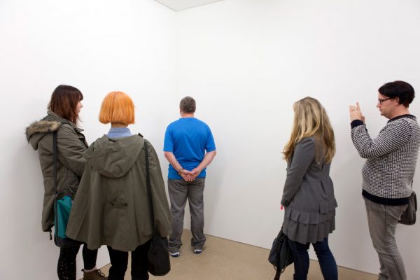 A middle-aged man in casual clothing stands, hands clasped behind his back, facing the corner of a bare, white room. Four people stand, watching the man.