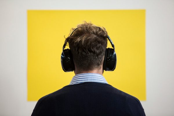 The back of a man’s head. The man is wearing large, black noise-cancelling headphones, looking towards a yellow-coloured square on a blank white wall.
