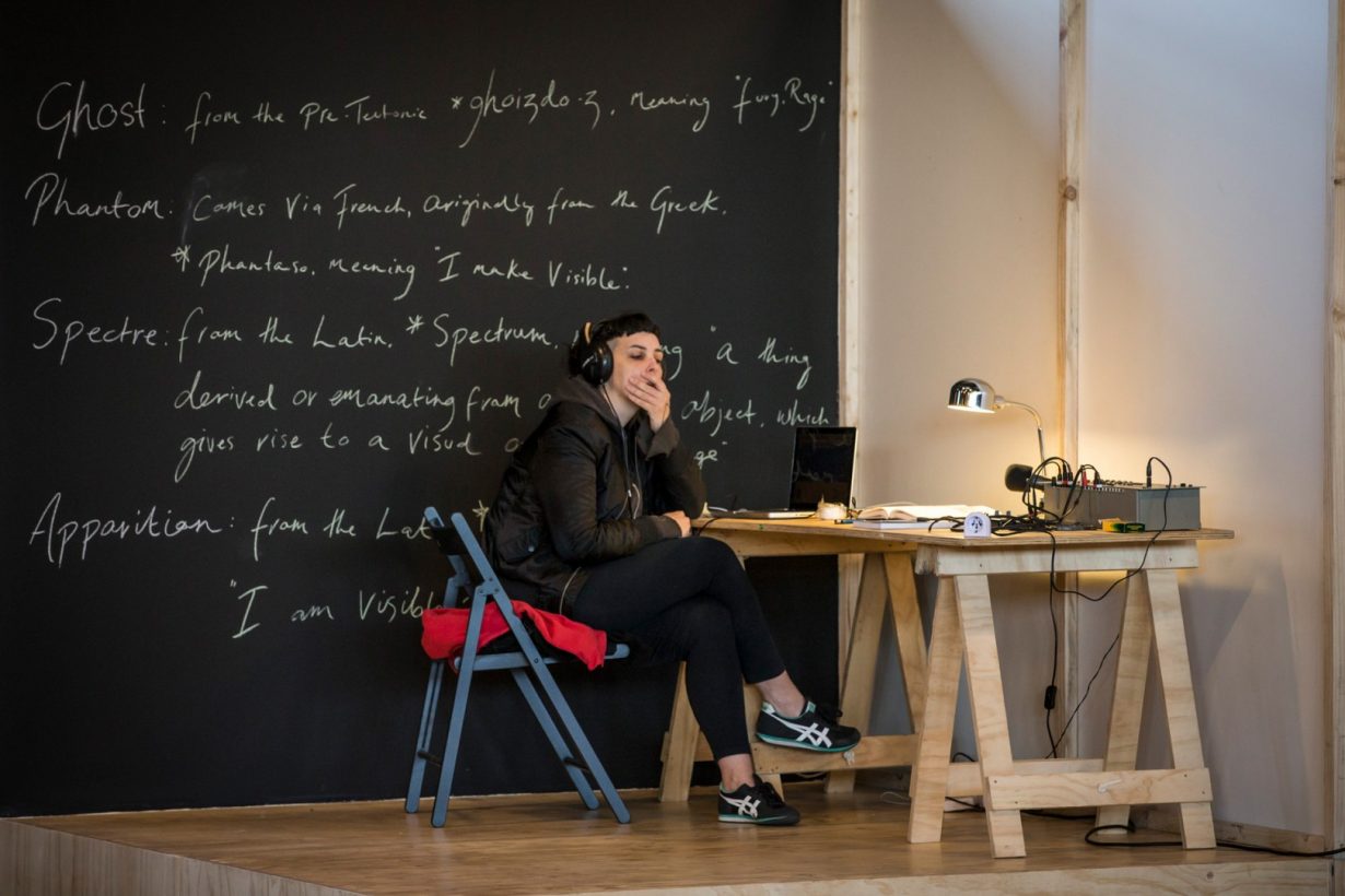 A person, wearing large headphones, seated at a basic plywood table, on top of a small plywood platform. On the table is a laptop, desk lamp, and electrical device connected with 4 audio cables. Behind the person is a large blackboard, with text handwritten in white chalk, including, “Ghost, Spectre, Phantom, Apparition.”