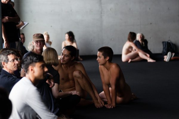 4 diverse dancers sit, naked, on the floor, in conversation with a fully clothed audience of around 12 people, sitting on the floor of a large, open space.