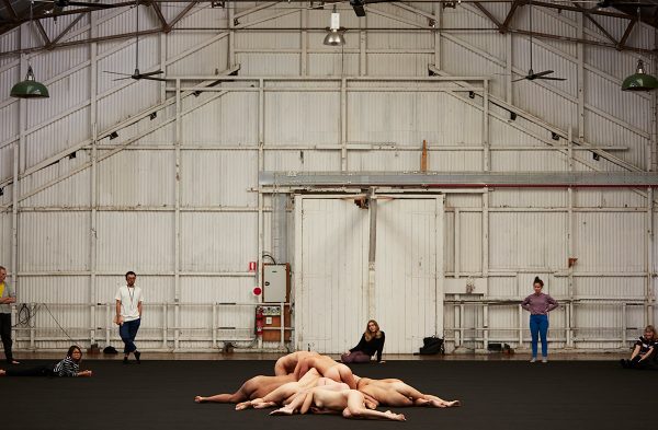 A diverse group of 9 dancers lies, with their naked bodies piled and overlapping on the floor, inside a large, open industrial space. Six people, dressed in casual clothing, stand and sit nearby, watching the dancers.