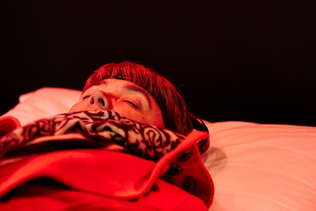A woman in a winter coat, wearing a simple black device around her forehead, lying on a white pillow, bathed in red light.