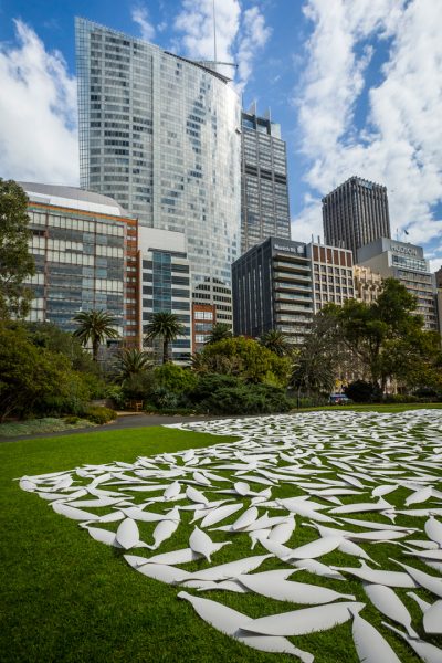 The lawn of an urban park, overlooked by tall office buildings. Hundreds of plain white shields are carefully arranged on the ground to form the corner of a geometric shape.