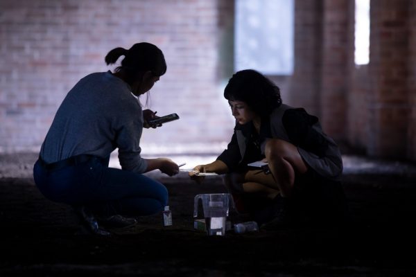 Two young woman crouch on the soil-covered floor of an old industrial building. The women are holding small notepads and a phone, and closely examining small squares of paper. In front of the women is a plastic jug of water and small squeeze bottle.