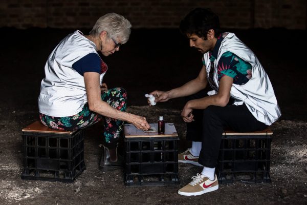 A young man and an older woman, both wearing silver reflective vests, sit on milk crates inside an old brick building. The floor of the space is entirely covered in soil. The man is tipping a small squeeze bottle over a small sample of soil on a milk crate.