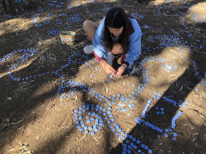 A teenaged girl in school uniform crouches, and places square blue tiles in a carefully arranged pattern of swirls on the ground.