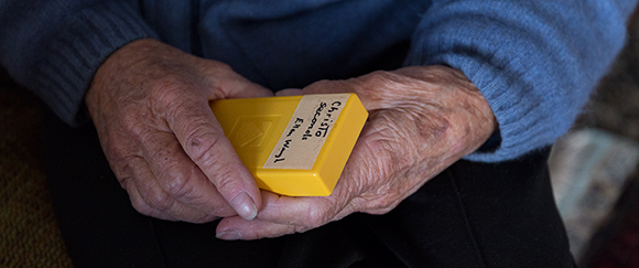 An elderly woman's hands, holding a small yellow plastic box.