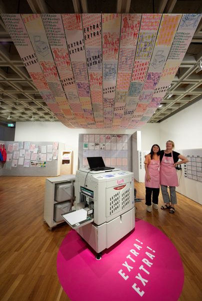 A large printing machine sits in the middle of an art gallery space. Two young women in identical pink aprons stand by the machine, arms around each other and smiling. The walls and floor of the gallery are decorated with coloured prints on paper, and a canopy of coloured prints is suspended from the ceiling.
