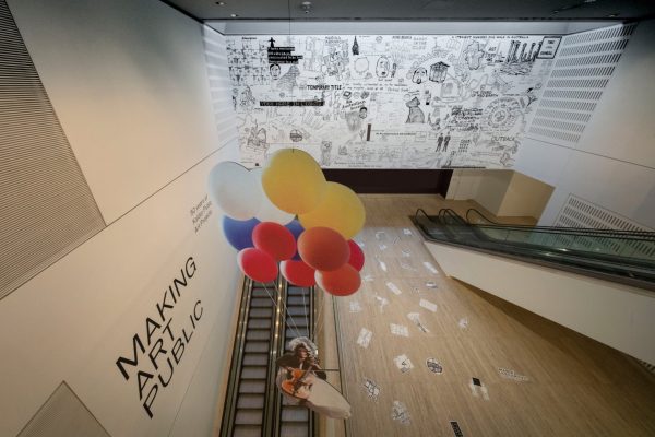 A cardboard cut-out of a woman playing a cello, suspended by coloured balloons, hanging in a large art gallery. On the rear wall of the gallery is a large, detailed hand-drawn sketch. On the side wall, large black text reads, “Making art public.”