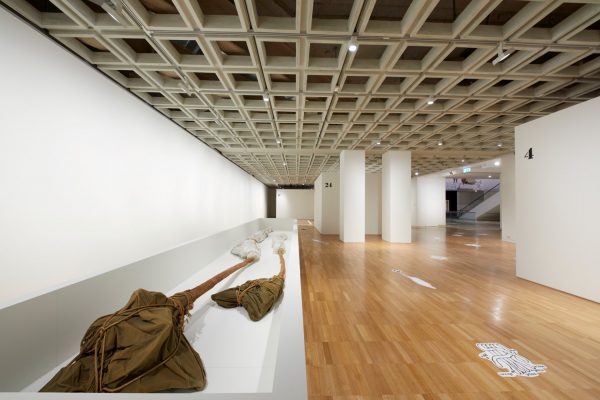 Two long, slender trees, with roots wrapped in khaki canvas and rope, and branches wrapped in plastic, lie on a white plinth in a large art gallery.