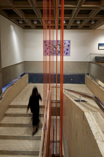 Around 20 long strands of dark orange rope, suspended vertically from the ceiling of an art gallery, and through the middle of a staircase.