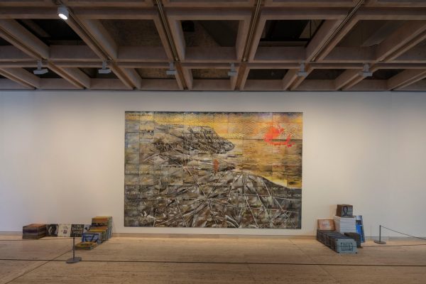 A large painting on the wall of an art gallery. The painting shows coastal cliffs wrapped in white fabric and rope, overlaid with text and chevron patterns. Stacks of small canvas boards sit on either side of the painting.