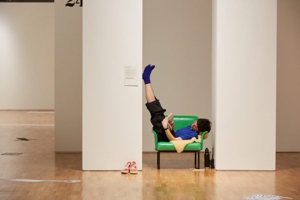 A young person lies, reading a book, on a small sheepskin rug on a green vinyl chair, inside an art gallery. The person wears socks, and rests their feet against a column.