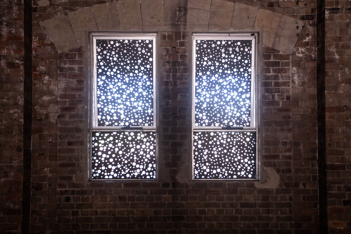 Two windows in an old, brick industrial building. The glass of the windows covered in black vinyl, with hundreds of small perforated holes, arranged in a pattern.