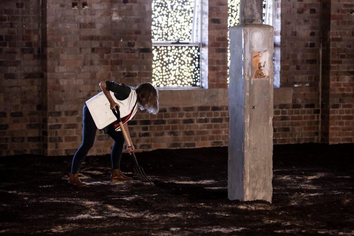 A woman, wearing a silver reflective vest, digs with a pitchfork into soil, which is laid across the floor of an old, brick industrial building.