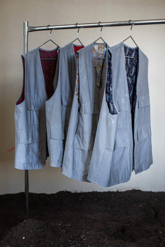 Four silver reflective vests, lined with 1970s patterned fabrics, hang on coathangers on a clothes rack inside a bare room. The floor of the space is entirely covered in soil.