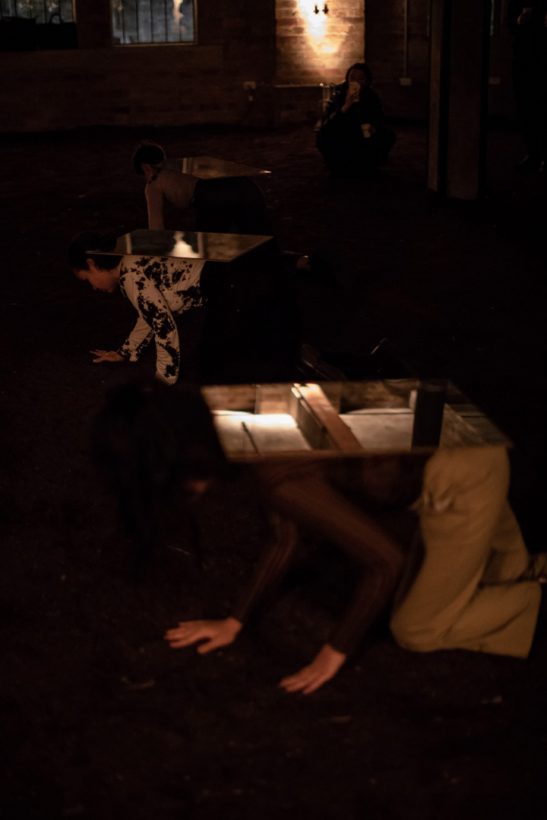 Three young women crawl across the soil-covered floor of an old, brick industrial building. The women carry mirrors on their backs.