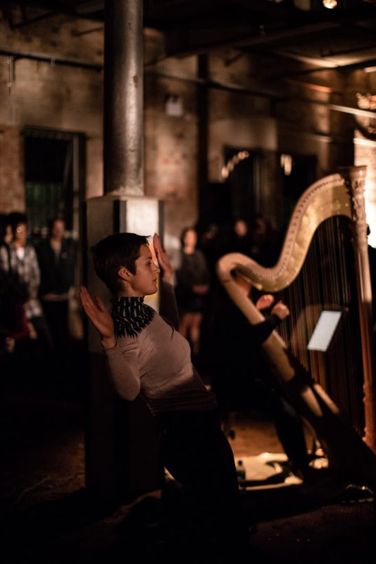 A young woman dances in an old, brick industrial building. A second young woman plays a harp. An audience of around 20 people stands in the darkened space, watching the woman.
