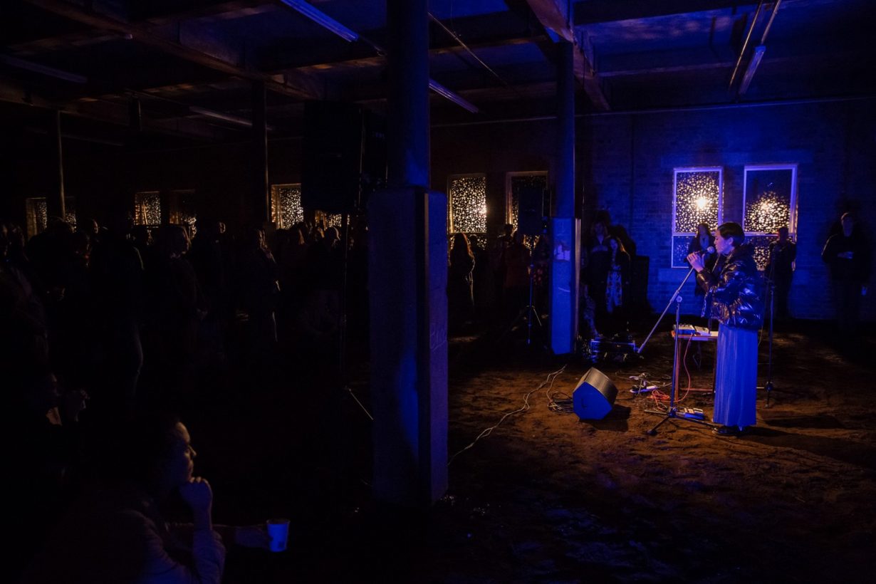 A young woman sings into a microphone, lit by blue light, in a darkened, old industrial building. The floor of the space is entirely covered in soil. A small audience of around 60 people watches.