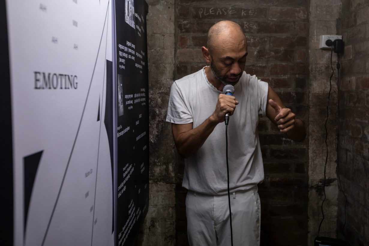 A man, dressed in white, speaking intently into a microphone, in a tiny, old brick room. On the wall are black-and-white graphic posters with excerpts of text.