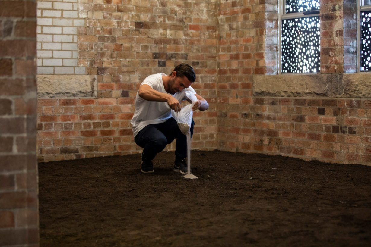 A young man crouches and pours a small bag of sand onto the soil-covered floor of an old industrial building.