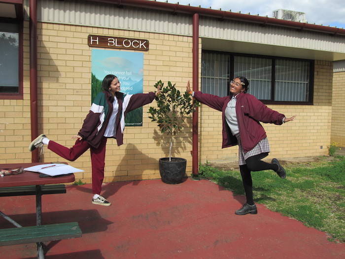 Two teenaged girls in school uniforms stand outside a brick building, with a sign reading "H Block". The girls are each smiling broadly, standing on one leg, and reaching towards each other with one hand, without touching. 