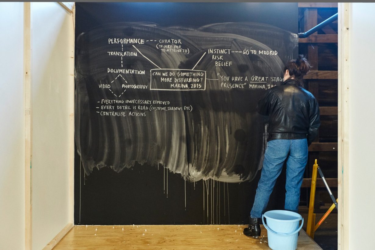 A young woman stands on a plywood platform, and creates a written diagram with white chalk on a blackboard. The blackboard is partly covered with large, messy white strokes and drips. The diagram includes the text, “Can we do something more disturbing? Marina, 2015.”