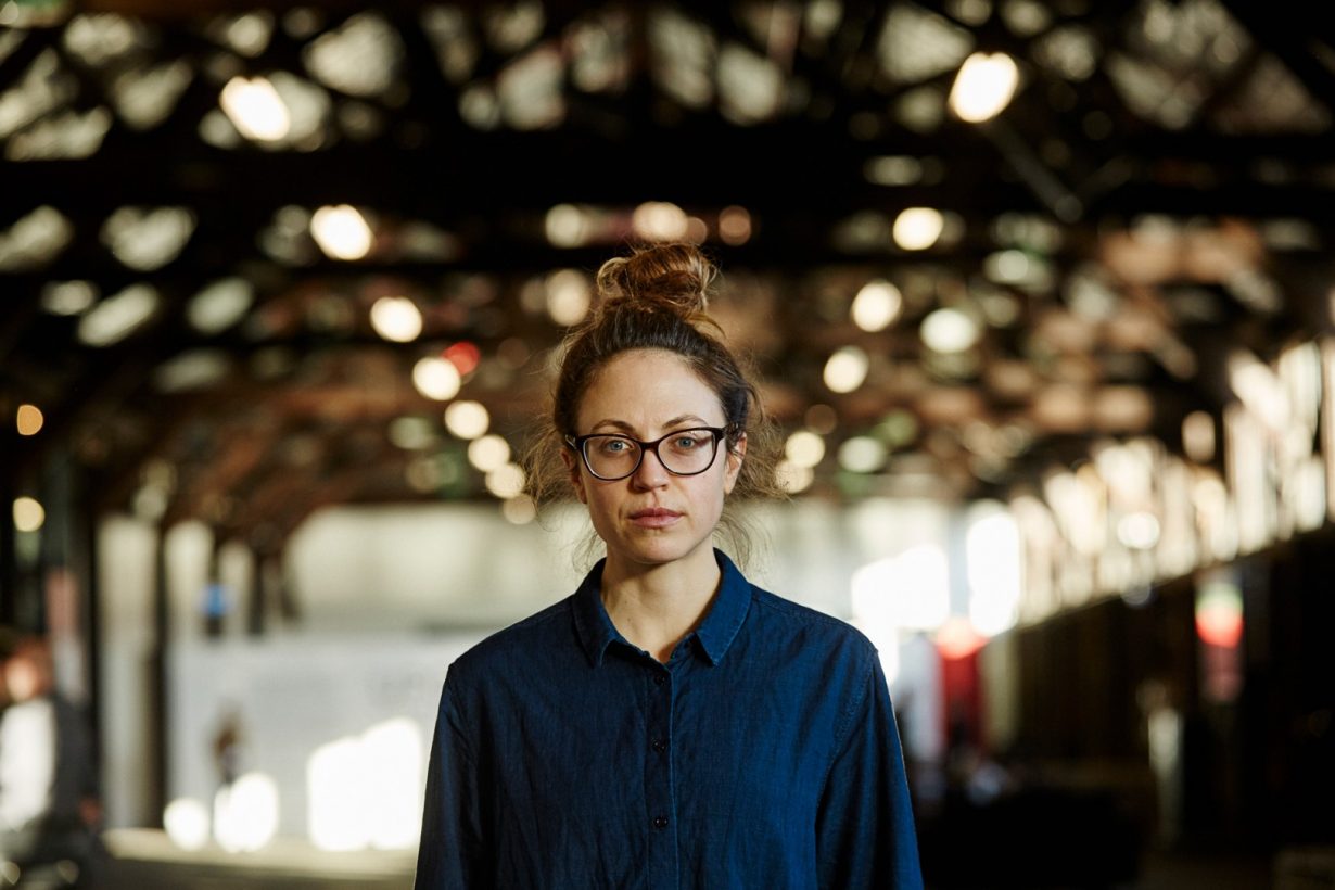 A young woman wearing glasses inside a large, open industrial building.