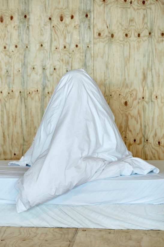 A mattress covered with a plain white sheet, inside a basic plywood structure. On top of the mattress is the shape of a person sitting under a pile of white bedclothes.