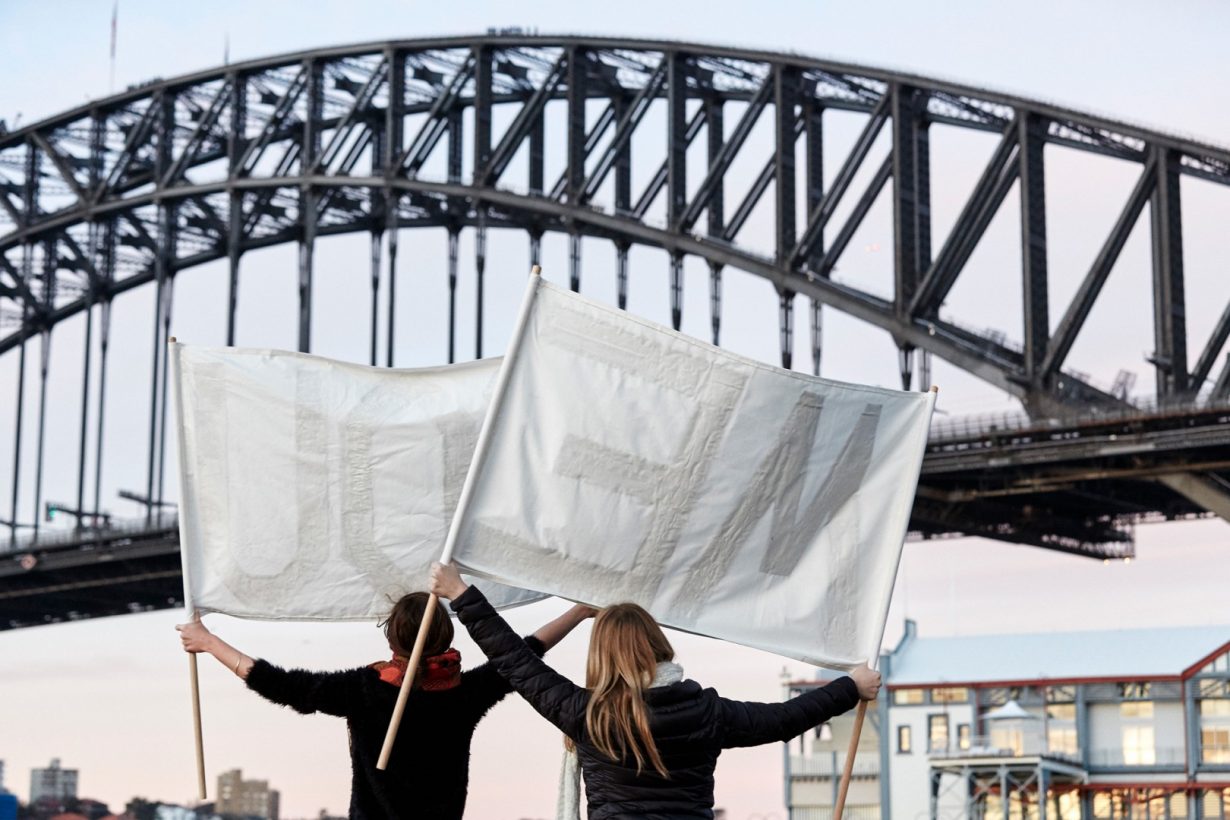 Two young women in winter clothing stand facing the Sydney Harbour Bridge, high above them. They are each holding up a simple white banner, seen from the back.