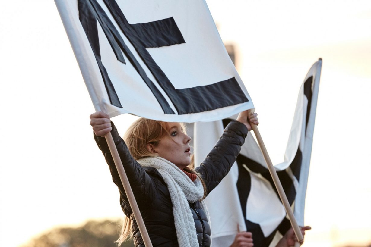 A young woman in winter clothing stands, holding up a simple white banner with large, plain black lettering in capital letters. The banner reads, “Me.”
