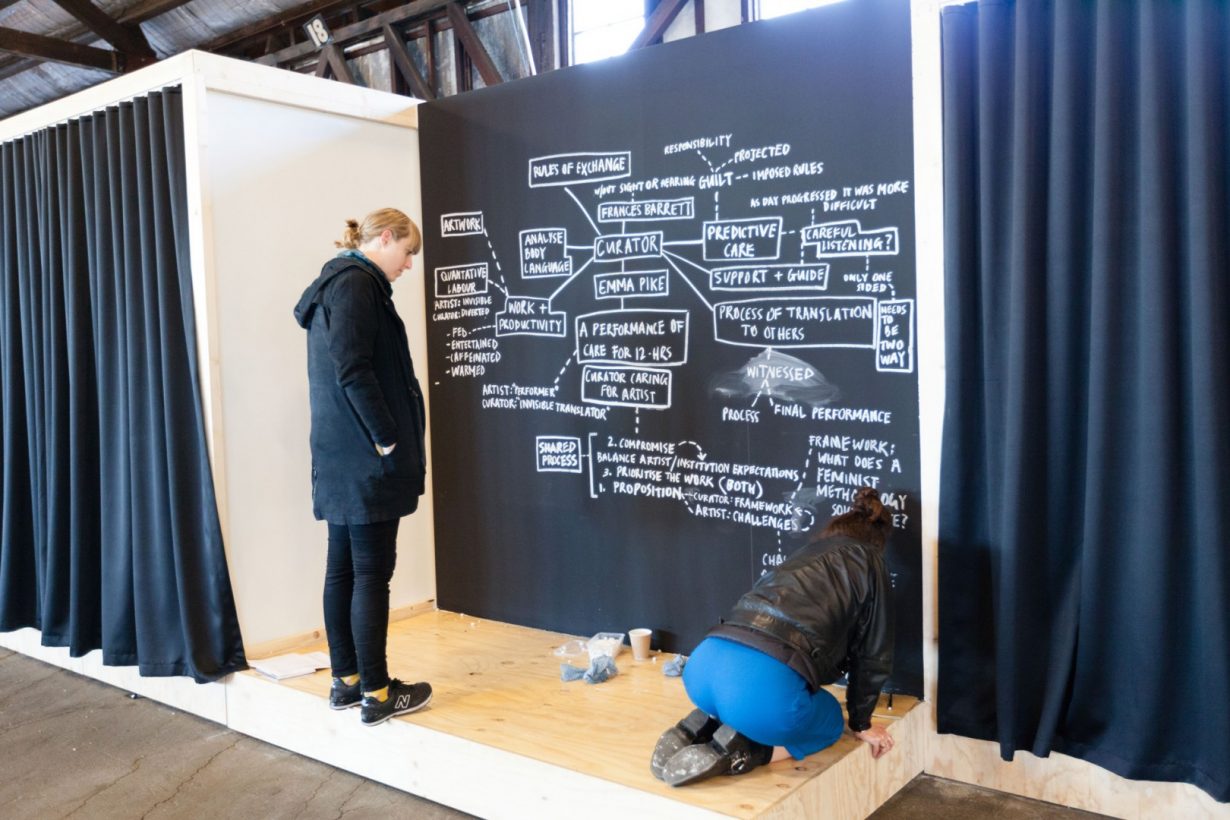 A young woman in a leather jacket kneels on a plywood platform, and creates a complex written diagram with white chalk on a blackboard. The diagram includes the text, “Curator, Emma Pike, A performance of care for 12 hours, Curator caring for artist.” A second woman in a winter coat stands on the plywood platform watching.