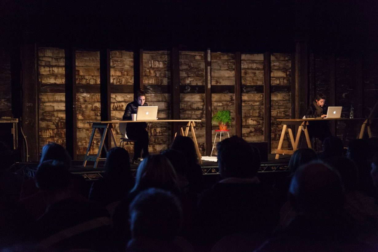 Two young women sit, dressed in winter coats, working at laptops, at two simple trestle tables in an old industrial building. The women are on a basic stage, lit by stage lighting. An audience sits in the darkened space, and watches the women onstage.