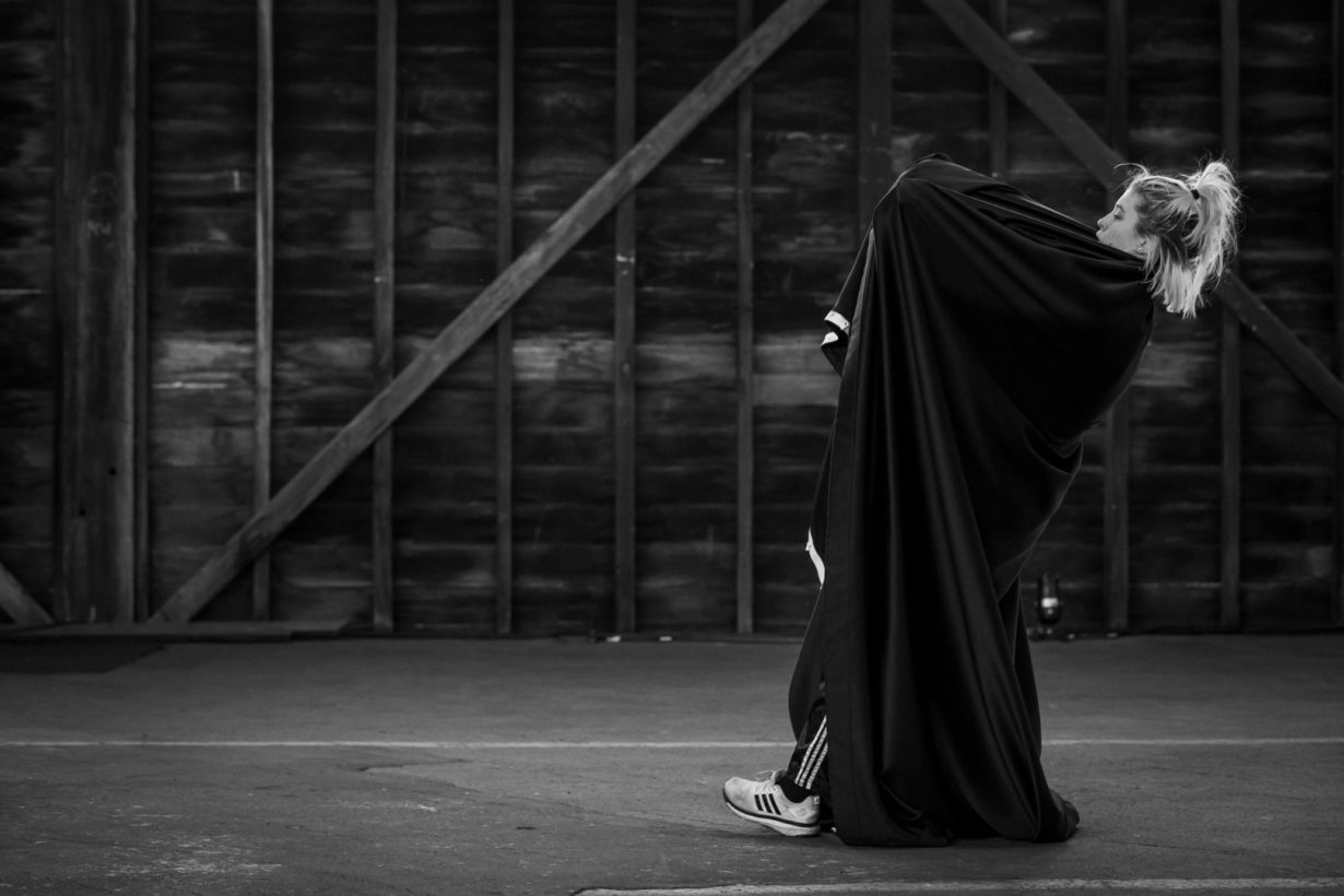 A young woman in Adidas sweat pants, wrapped in a large black cloth, leaning backwards and pulling on the cloth, in a large, open industrial building.