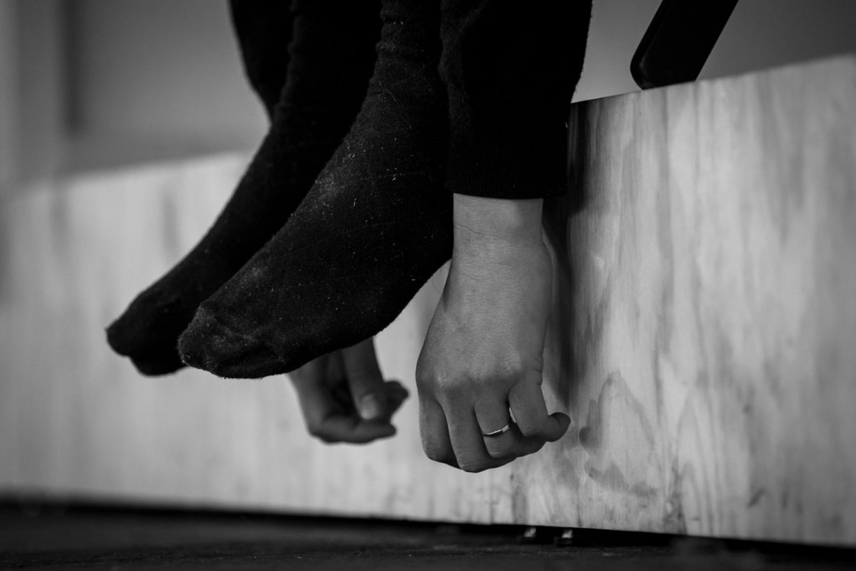 A young woman’s hands, wearing a simple wedding ring, and her feet, in black socks, hang over the edge of a basic plywood platform.