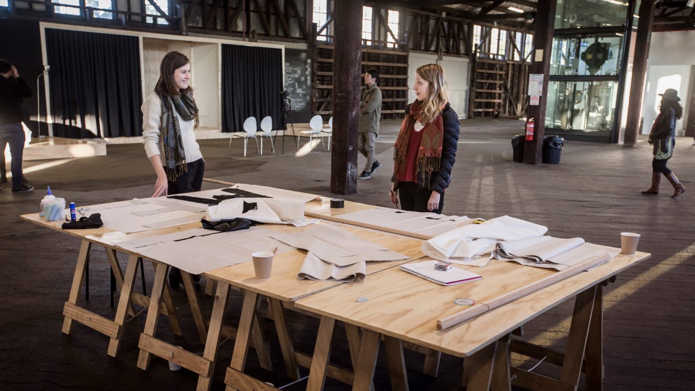 Two young women in winter clothing stand at a large workspace, which consists of four plywood tables pushed together, inside an old industrial building. The tables are littered with pieces of black and white fabric, tape and coffee cups. The women are looking at each other and smiling.