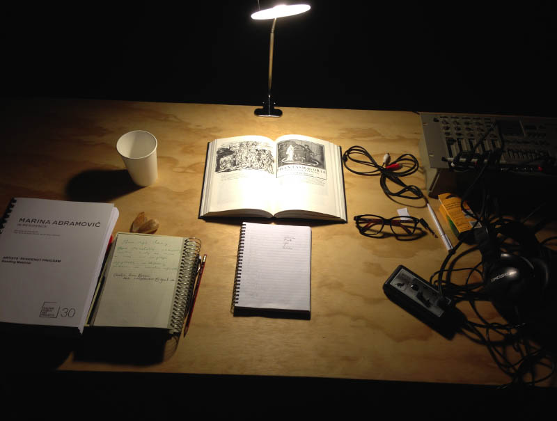 A basic timber desk, with a desk lamp. On the desk are arranged a spiral-bound Residency guide, two notebooks, pencil and pen, disposable paper cup, a pair of glasses, a sound desk, headphones, around 4 audio cables, and an open book with the text Phantasmagoria. 