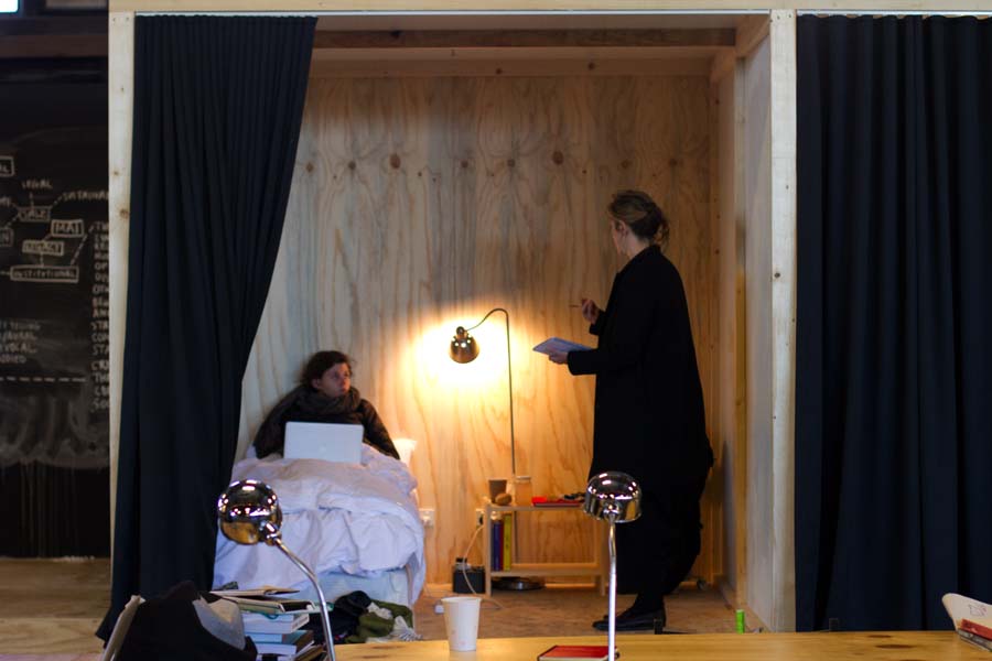 A young woman, wearing a winter coat and scarf, lies in a single bed with plain white bedsheets, inside a small plywood cubicle. The woman has a laptop computer on her lap, and is talking with a second woman, who stands in the cubicle, holding a pencil and notepad. 