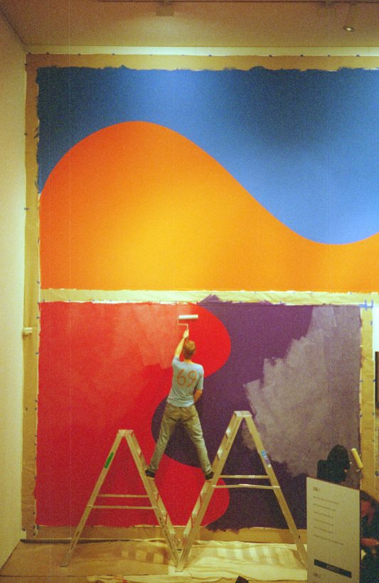 A young man stands, straddling two ladders, painting a geometric arrangement of orange blue, red and purple and curves, marked out with masking tape, on a tall art gallery wall.