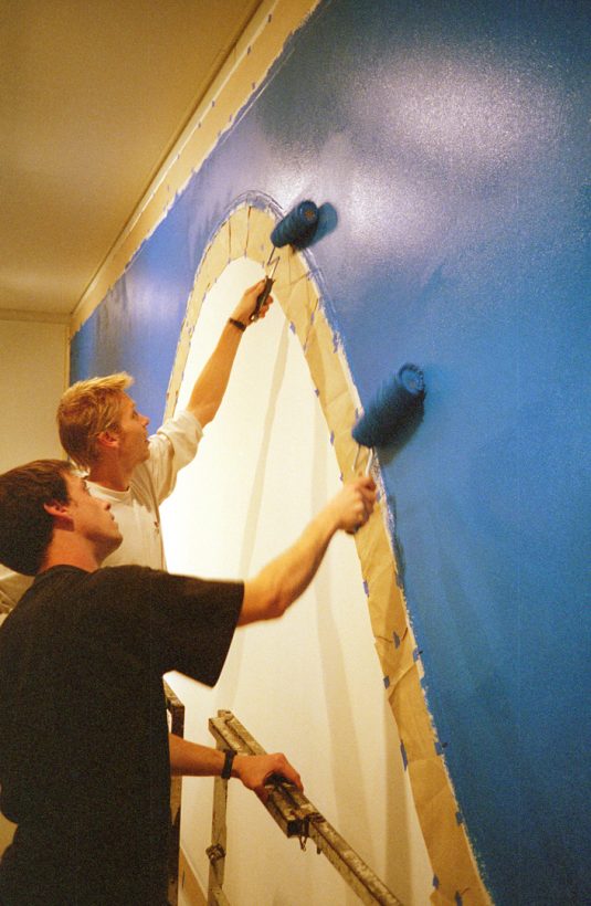 Two young men use rollers to paint a blue curve on an art gallery wall, marked out with masking tape.