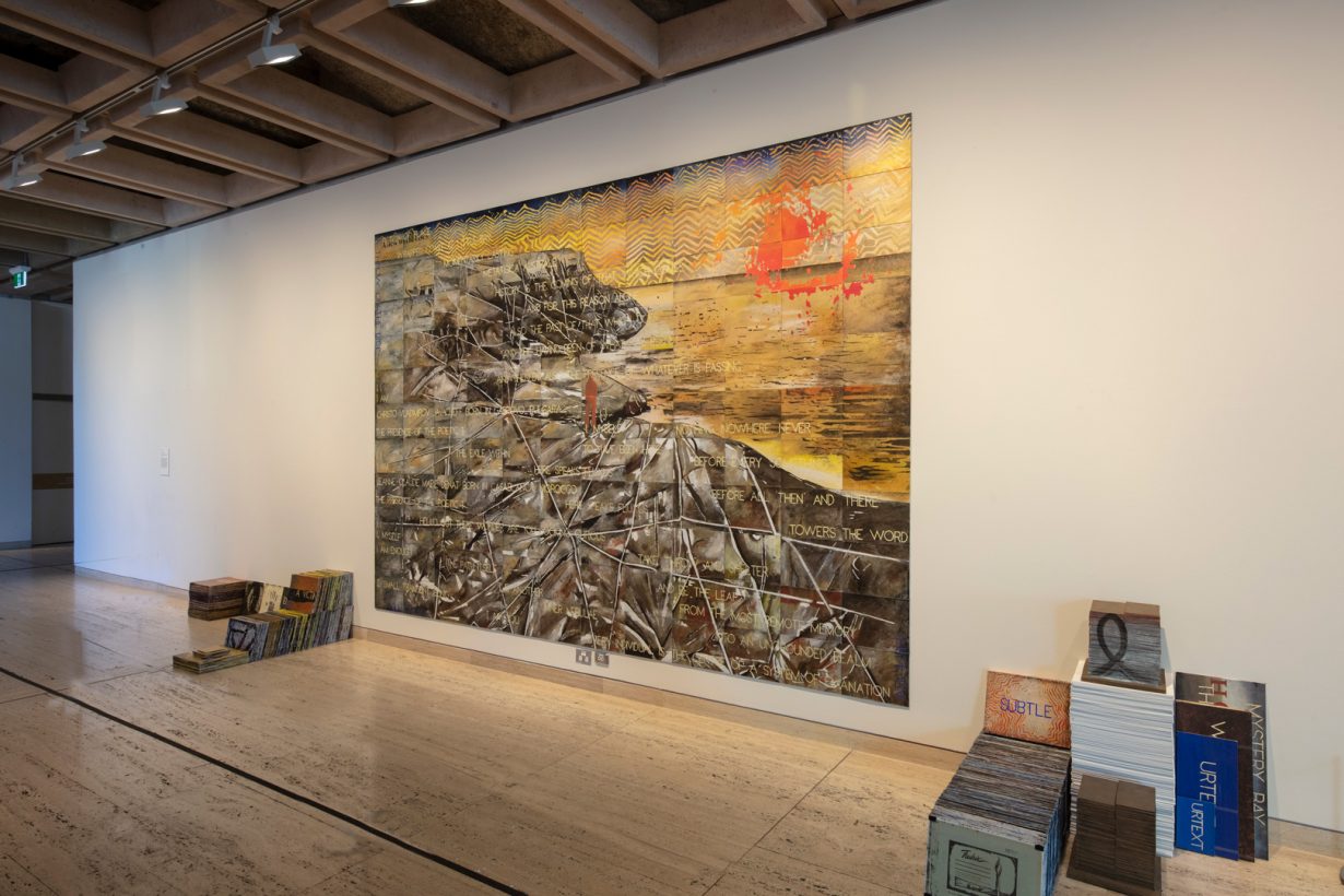 A large painting on the wall of an art gallery. The painting shows coastal cliffs wrapped in white fabric and rope, overlaid with text and chevron patterns. Stacks of small canvas boards sit on either side of the painting.
