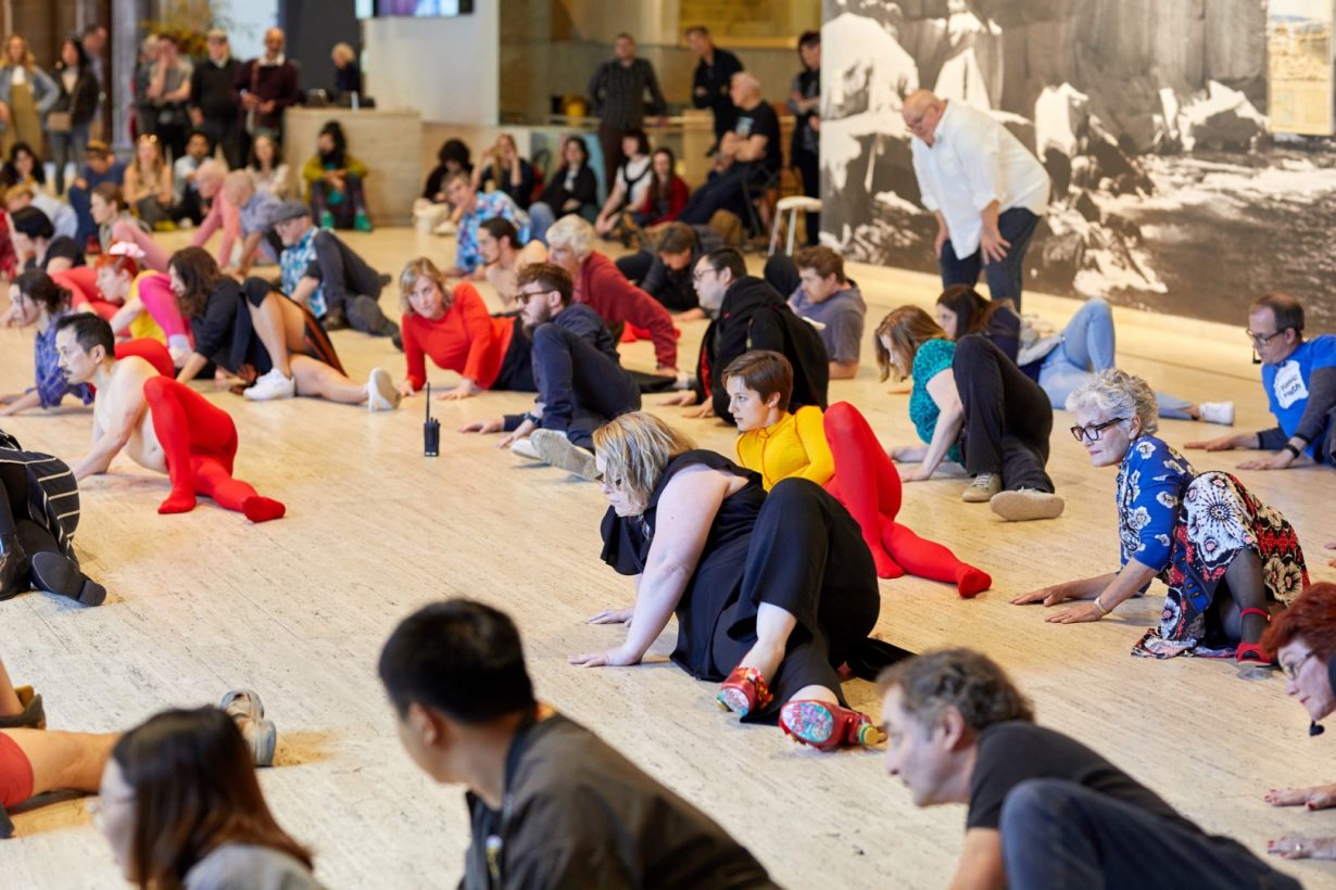 A group of around 40 diverse people lie on the floor of an art gallery. The people are lying on their sides, in an identical pose, with one knee bent upwards, and looking in the same direction, with serious expressions. An audience of around 40 people stands and sits on the edge of the group, watching.