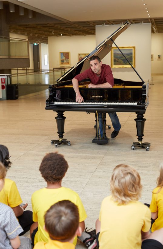 A man stands inside a grand piano, and leans forward to play the piano keys with one hand. The piano has wheeled legs and a round hole cut in the lid. A group of primary school children is seated on the floor, watching the pianist, inside an art gallery.