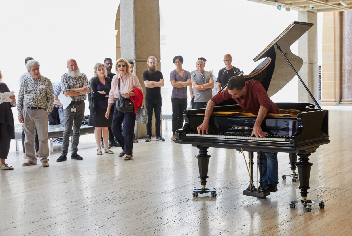 A man stands inside a grand piano, and leans forward to play the piano keys, with a look of intense concentration. The piano has wheeled legs and a round hole cut in the lid. A crowd of around 15 people is watching the pianist, inside a modern art gallery.