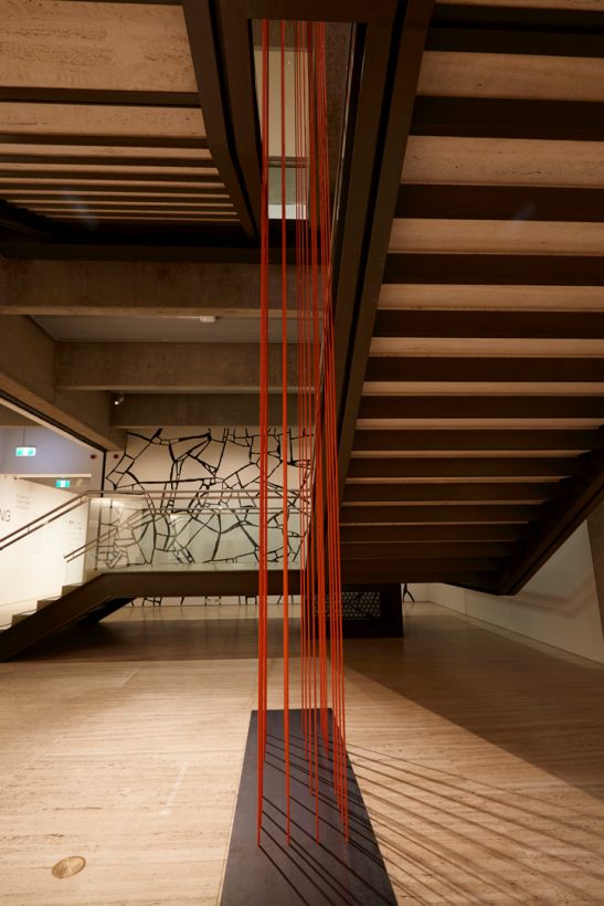 Around 20 long strands of dark orange rope, suspended vertically through the middle of a staircase in an art gallery, and attached to a heavy base on the floor.