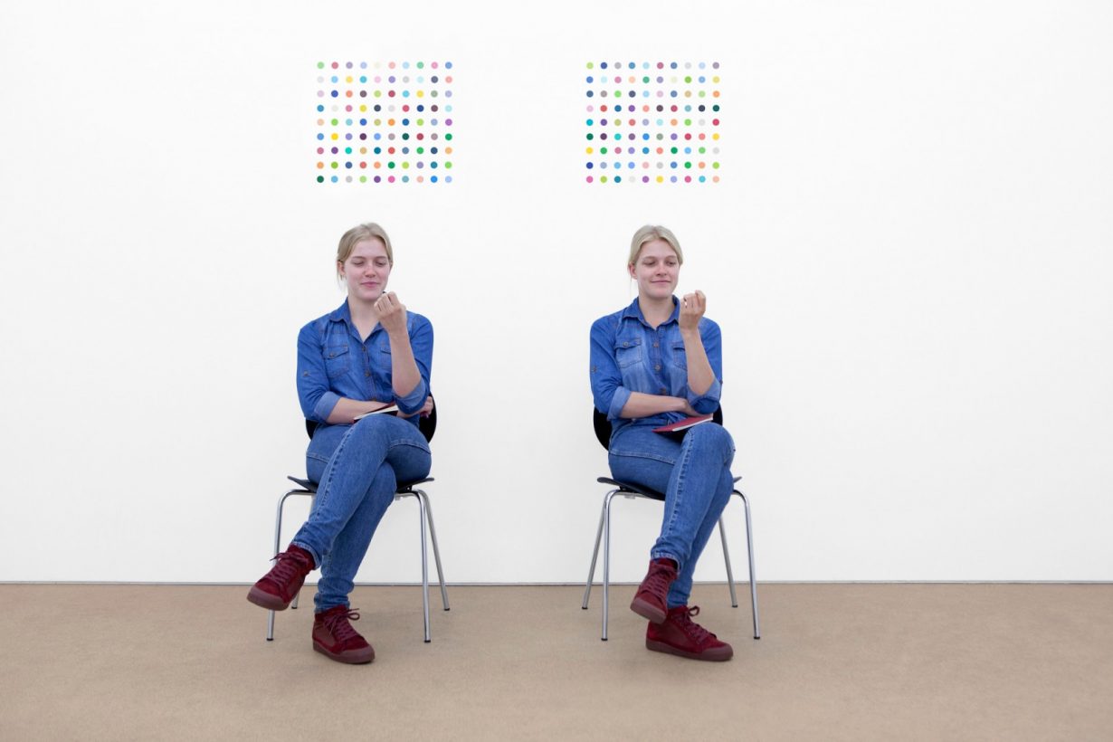 Two identical young women, dressed in identical denim shirts, jeans and red shoes, sit on identical chairs, holding identical books, and gaze at their fingernails with amused expressions. On the bare white wall behind the women are two similar paintings, consisting of grids of brightly coloured dots.