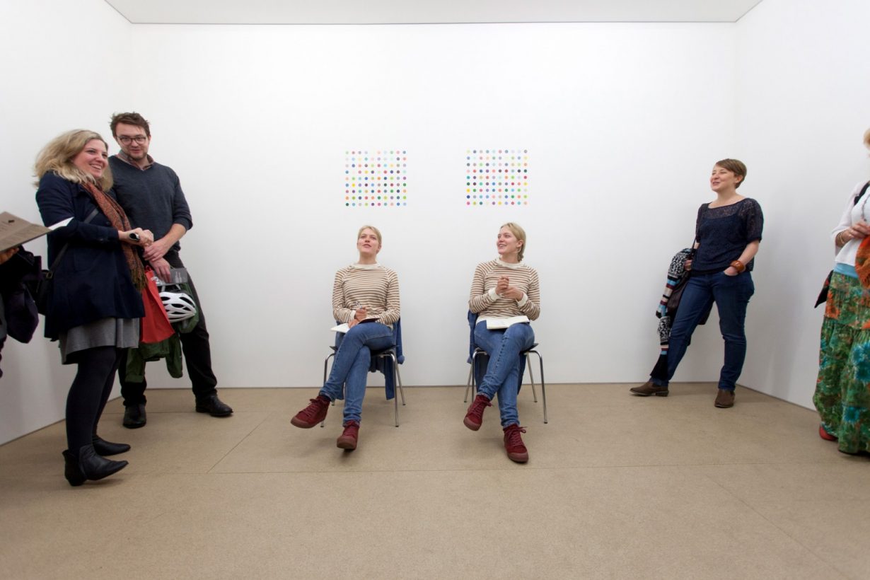 Two identical young women, dressed in identical jumpers, jeans and red shoes, sit on identical chairs, holding identical books and smiling. On the bare white wall behind the women are two similar paintings, consisting of grids of brightly coloured dots. 5 people stand, watching, smiling and conversing with the young women.